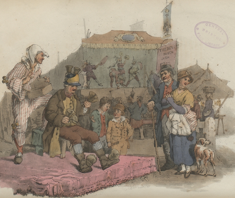 A man sits on a red carpet with bells on his hat and shoes, and with three further bells in his hands.  A man in a white hat and reda nd white striped suit stands behind him.  A couple have stopped to watch the performance, and a fair can be seen in the background.